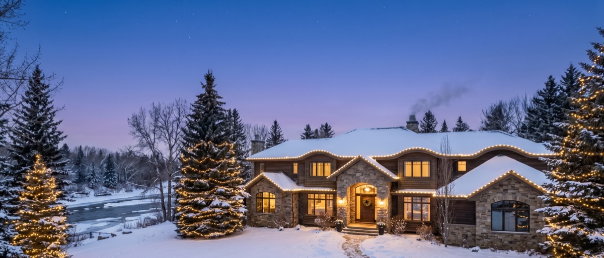 A large riverfront estate home in Elbow Park, Calgary with a long roofline elegantly lit in warm-white lights and mature trees wrapped in lights at dusk.
