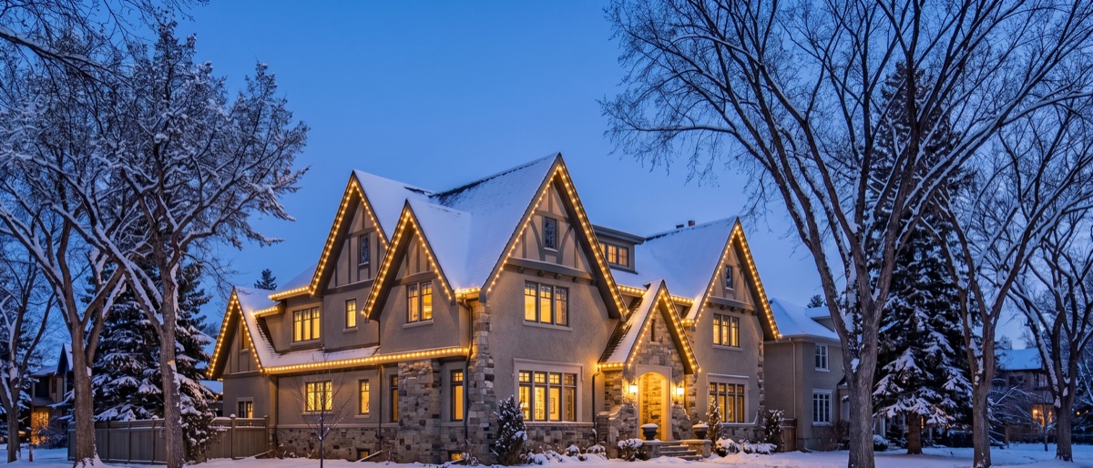 A heritage Mount Royal home in Calgary with steep gables outlined in warm-white Christmas lights on a snowy evening.